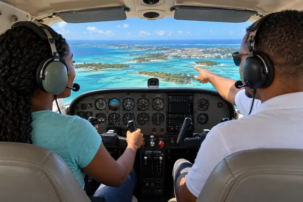 Student at the controls of a small airplane beside an instructor, with turquoise coastal waters visible ahead.